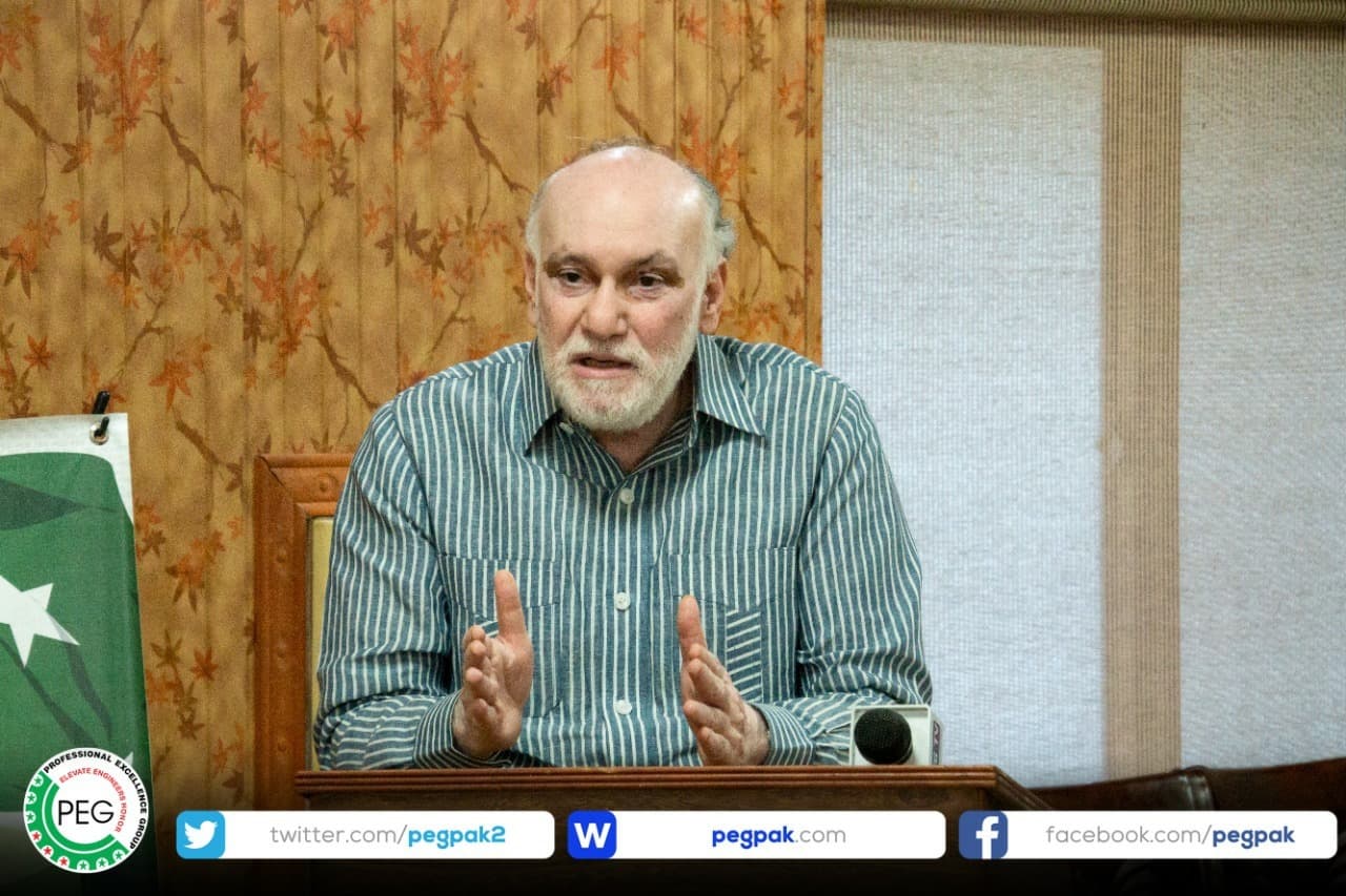 Engr. Waseem Nazir speaking at a microphone with the Pakistani flag behind him at Mangla Guest House.